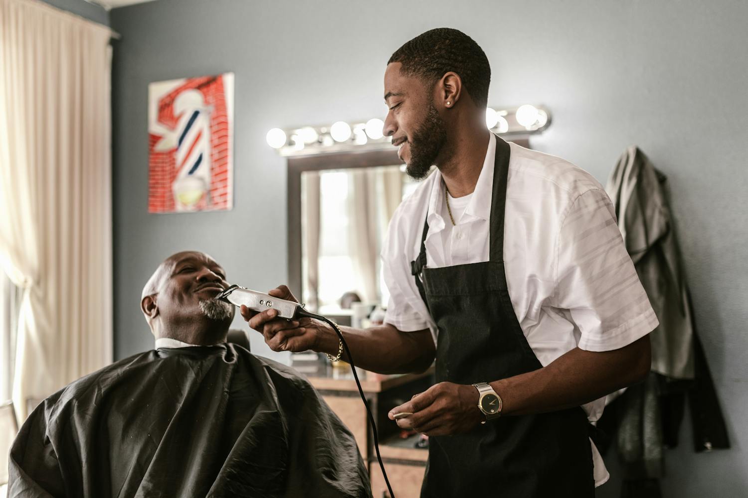 A barber smiling while detailing an older client's beard with clippers in the boutique