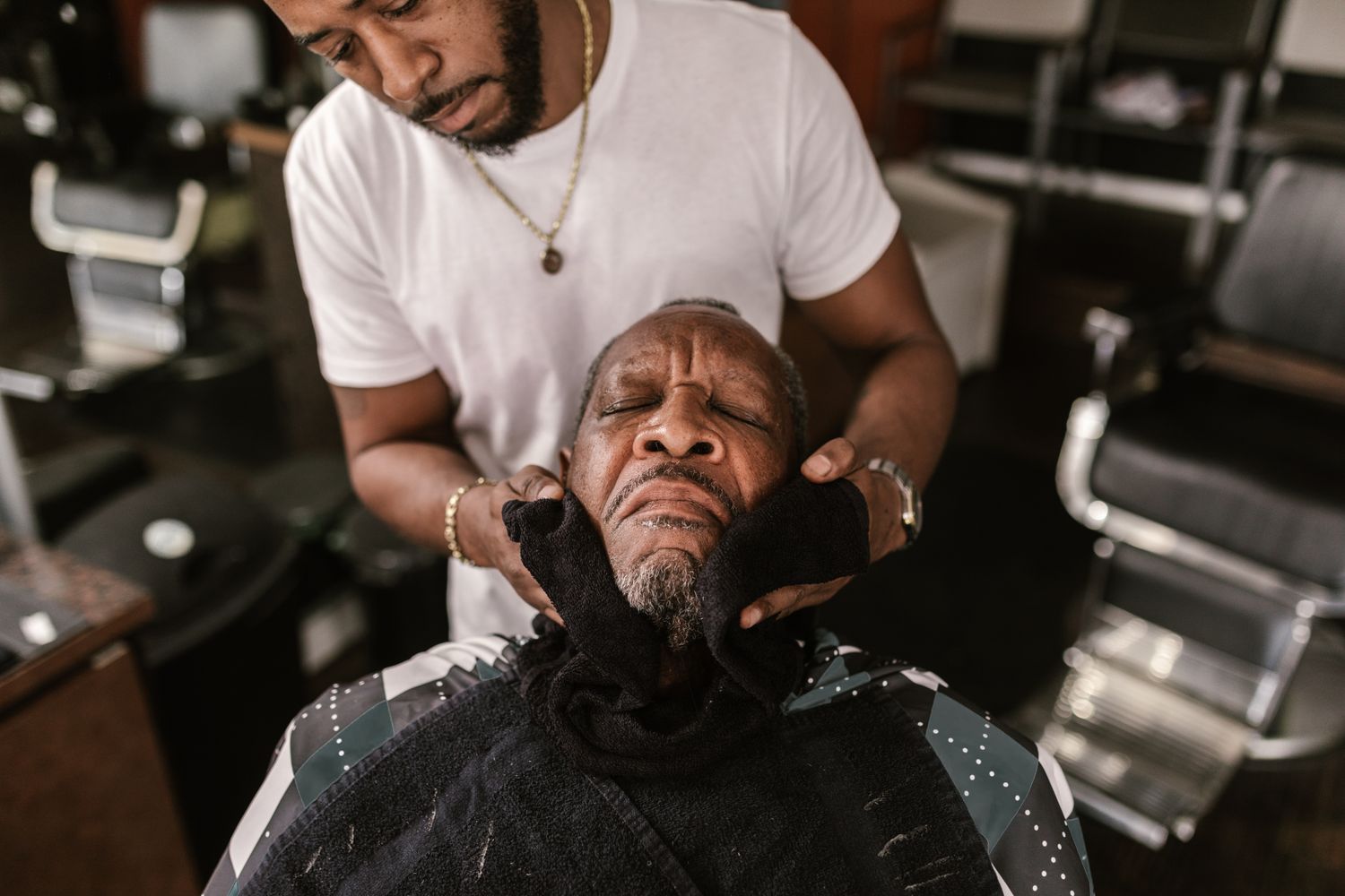 A barber finishing a client's grooming with a warm towel, the client relaxed with eyes closed